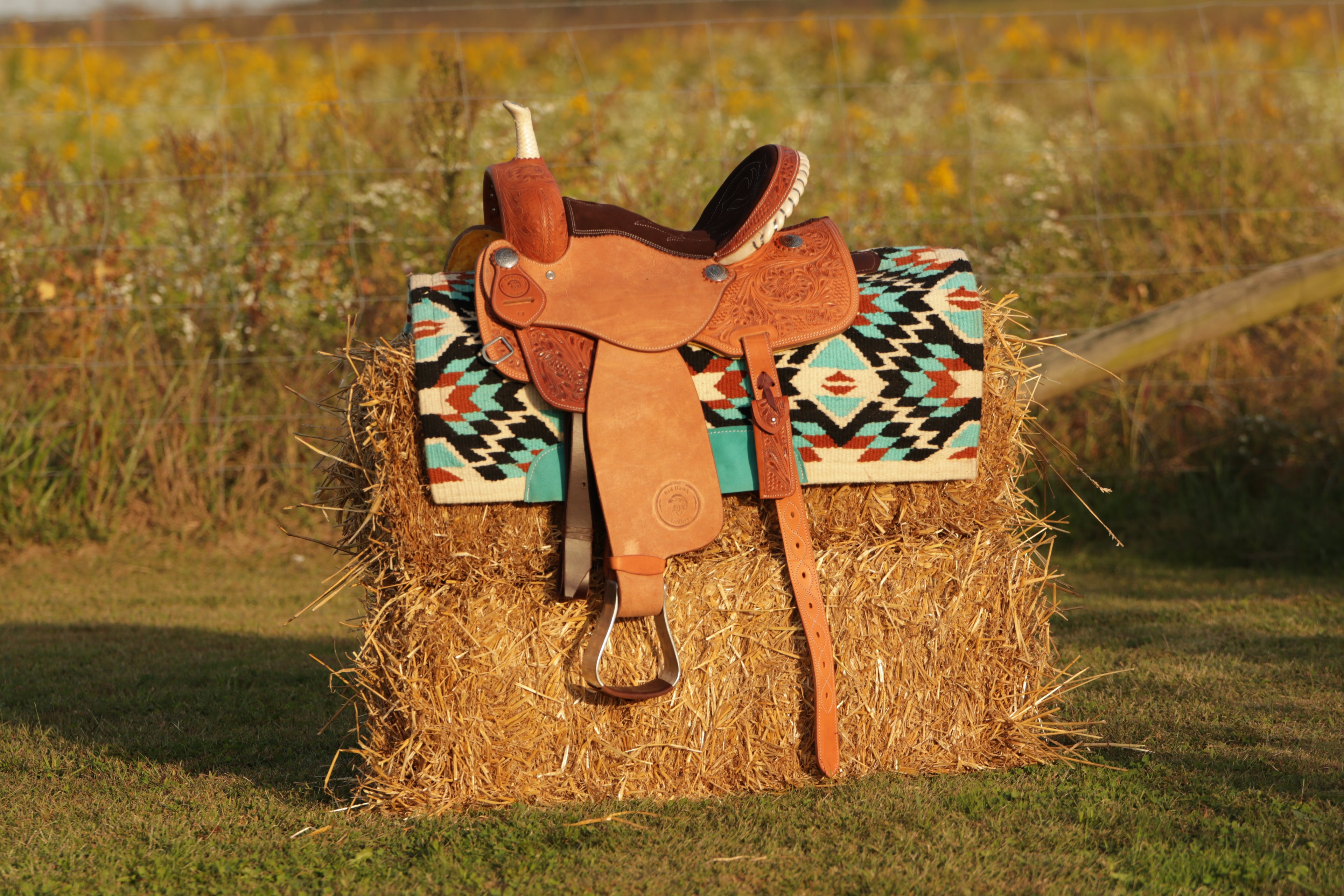 Brown leather saddle on a hay bale with a colorful blanket in a field